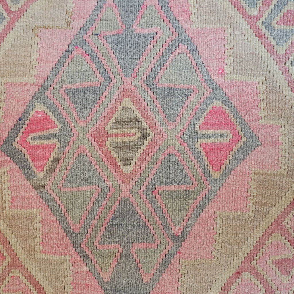 A close-up of a woven textile with a geometric kilim-style pattern in pink, beige, and gray.
