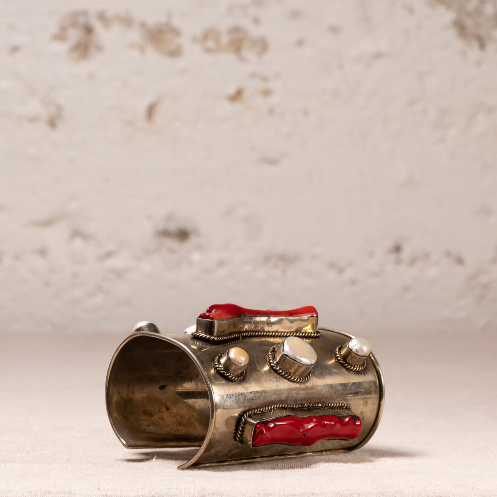 A vintage-style silver-toned cuff bracelet with red coral inlays and white pearl accents, displayed on a light beige textured surface against a rustic, light-colored stone wall.