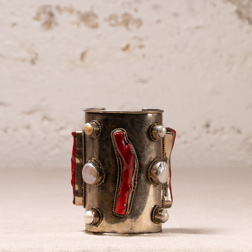 A vintage-style silver-toned cuff bracelet decorated with red enamel inlays, white pearl-like beads, and small silver studs, set against a textured off-white background.