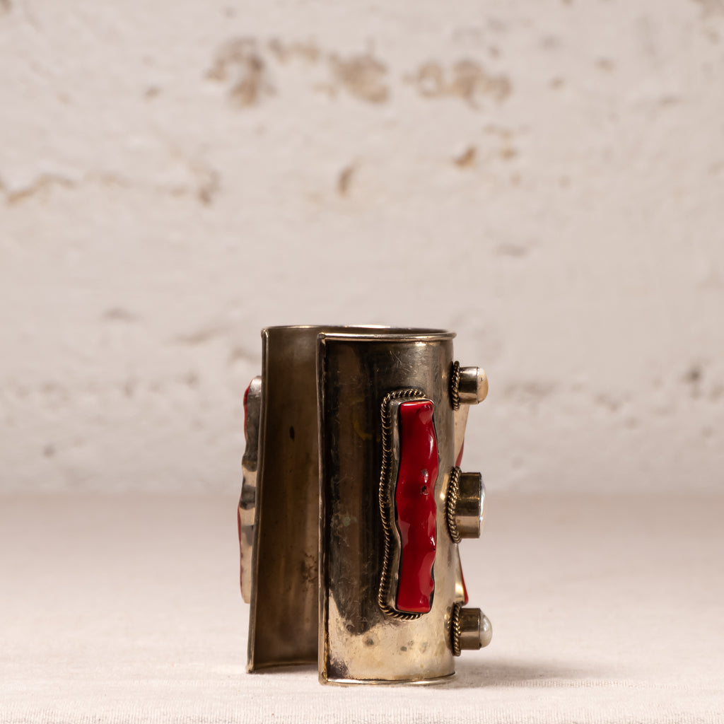 A vintage silver-toned cuff bracelet with red inlay details, standing upright on a light beige textured surface against a matching light beige background.