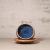 A handmade ceramic bowl with a vibrant blue glaze inside and a rustic terracotta-colored exterior, shown resting on a matching saucer against a textured off-white plaster wall.