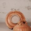 A rustic handmade ceramic lidded pot with a matching saucer, featuring a warm terracotta orange-brown glaze and a speckled beige center, set against a textured off-white wall.
