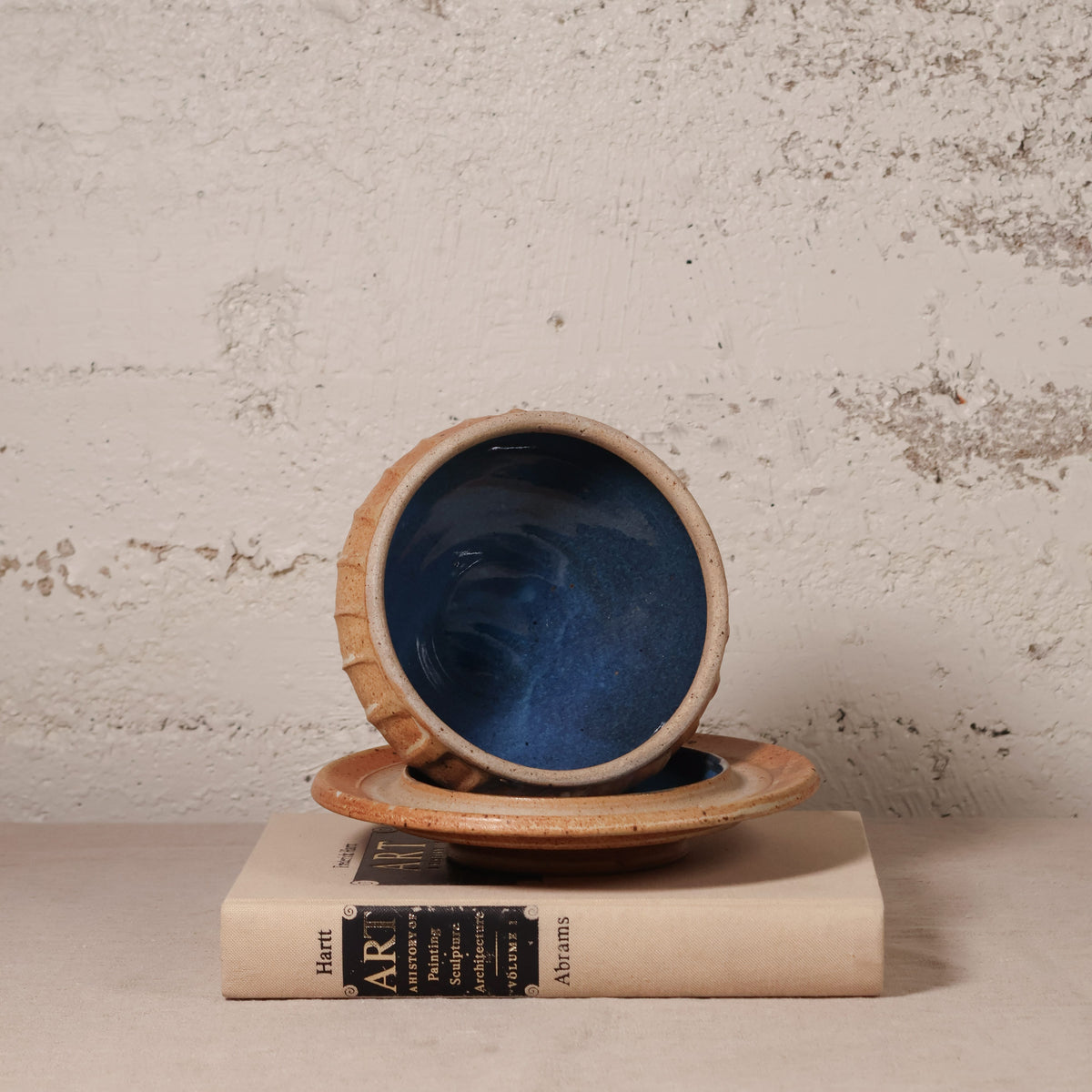 A handmade ceramic bowl with a deep blue interior and a textured beige exterior, shown resting on a matching saucer and a vintage book against a rustic plaster wall.