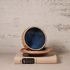 A handmade ceramic bowl with a deep blue interior and a textured beige exterior, shown resting on a matching saucer and a vintage book against a rustic plaster wall.