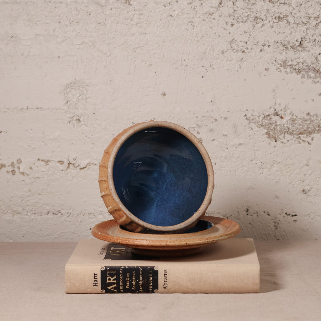 A handmade ceramic bowl with a deep blue interior and a textured beige exterior, shown resting on a matching saucer and a vintage book against a rustic plaster wall.