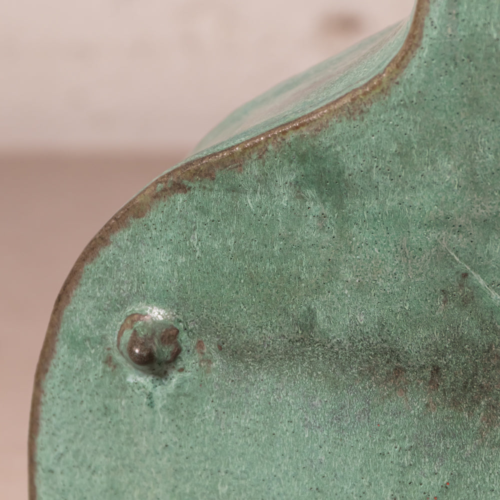 Close-up of a green metal surface with a weathered patina and a small rusted screw detail, showing part of a curved object against a soft beige background.