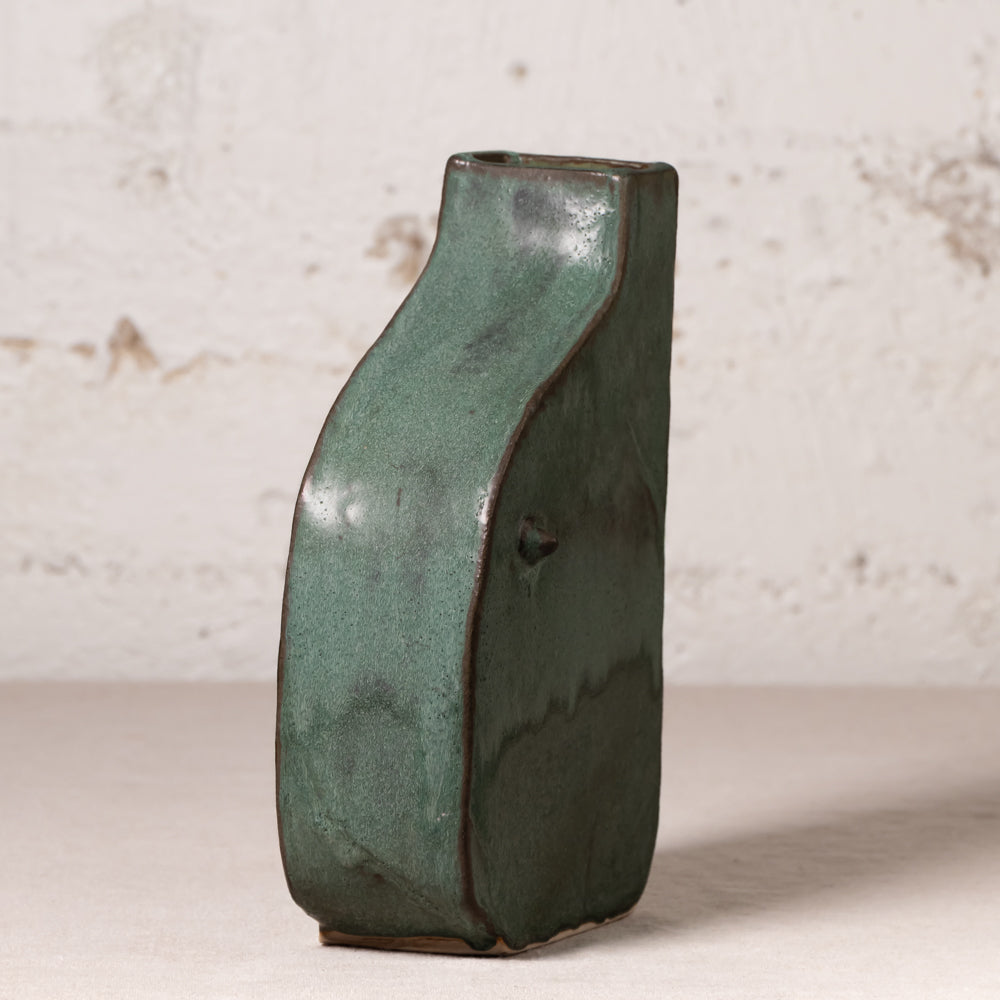 A rustic handmade ceramic vase with a wavy silhouette and a textured dark green glaze, standing on a light surface against a distressed off-white wall.