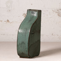 A rustic handmade ceramic vase with a wavy silhouette and a textured dark green glaze, standing on a light surface against a distressed off-white wall.