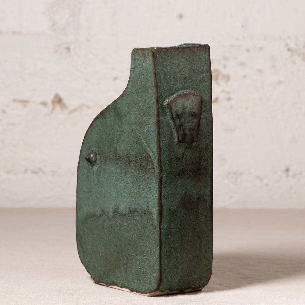 A handmade ceramic vase with a dark green glaze and a sculpted dog head detail on the front, set against a rustic white brick wall background.
