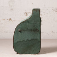 A small sculptural ceramic vase with a dark green glaze and a small protruding knob, photographed against a light textured wall.