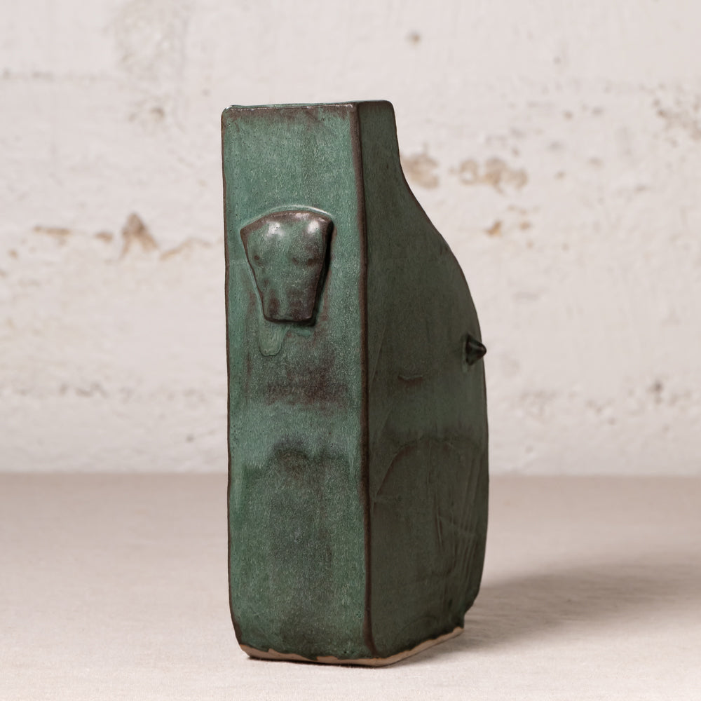 A handmade ceramic vase with a textured, rustic finish in dark green glaze, featuring a sculpted animal head detail on the side, set against a light textured wall.