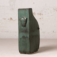 A handmade ceramic vase with a textured, rustic finish in dark green glaze, featuring a sculpted animal head detail on the side, set against a light textured wall.