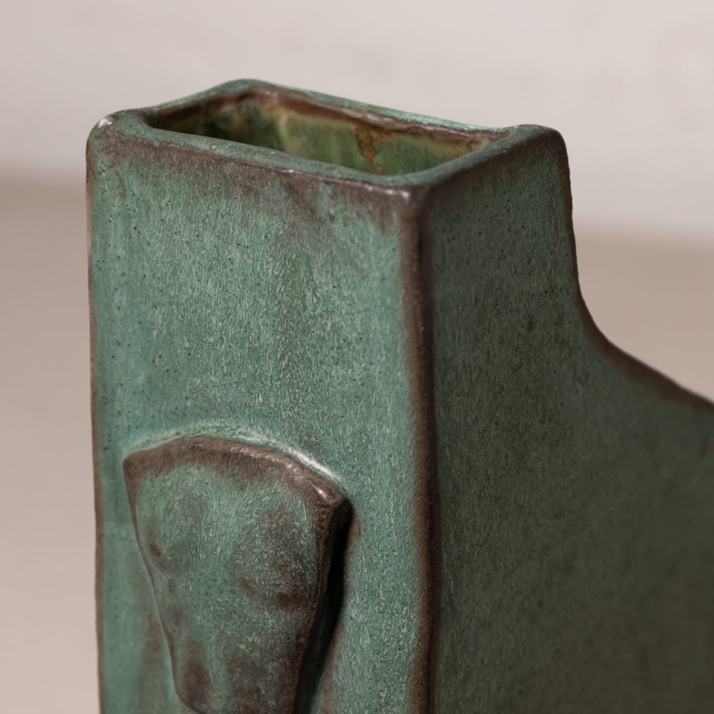 Close-up of a square ceramic vase with a textured sage green glaze and dark brown unglazed edges, showing a small raised decorative detail on the front.