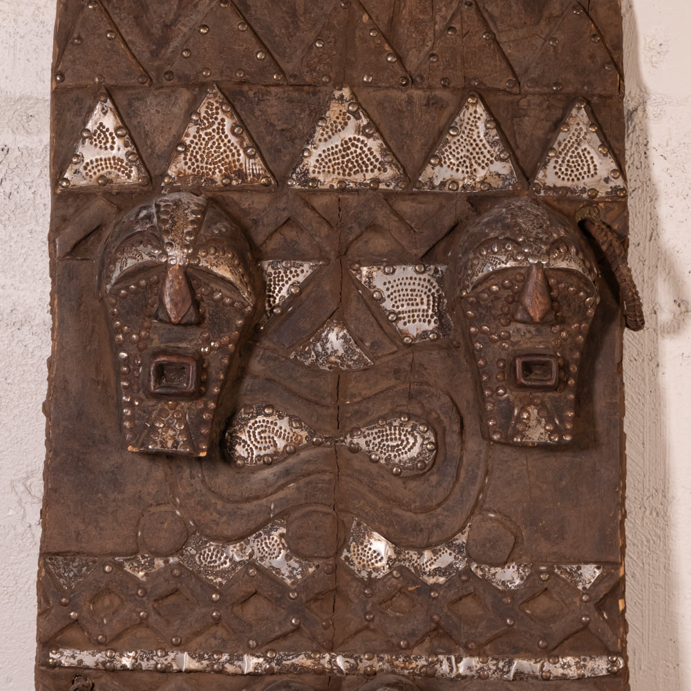 An antique carved wooden African wall plaque featuring two stylized faces and geometric patterns, decorated with silver-painted metallic accents.