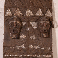 An antique carved wooden African wall plaque featuring two stylized faces and geometric patterns, decorated with silver-painted metallic accents.