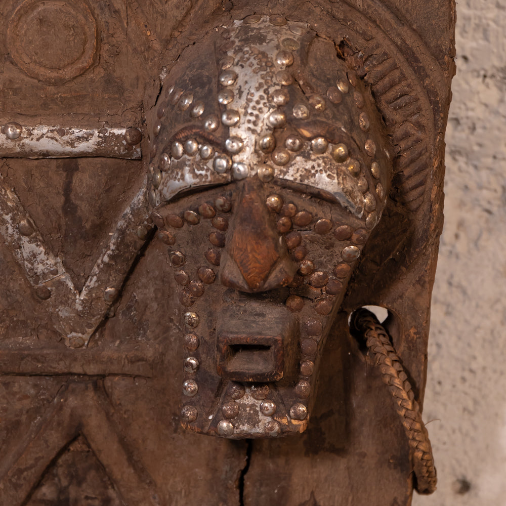Close-up of an antique carved wooden door featuring a detailed metal lion head mask with raised studs, a hanging rope, and weathered brown wood tones against a light textured wall.