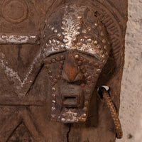 Close-up of an antique carved wooden door featuring a detailed metal lion head mask with raised studs, a hanging rope, and weathered brown wood tones against a light textured wall.