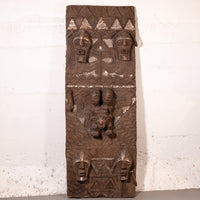 A vertically oriented carved wooden wall plaque with intricate tribal-style relief carvings and geometric patterns, shown leaning against a white textured wall.