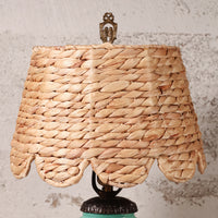 A woven natural fiber lampshade with a scalloped edge and a decorative metal finial on top, shown against a textured light gray wall.