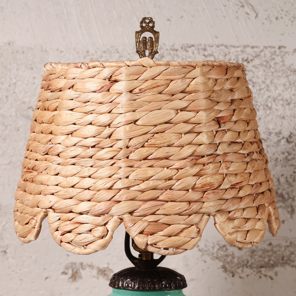 A woven natural fiber lampshade with a scalloped edge and a decorative metal finial on top, shown against a textured light gray wall.