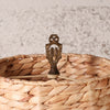A close-up of a woven natural fiber basket with a decorative bronze-toned metal finial on top, set against a softly blurred light gray textured background.
