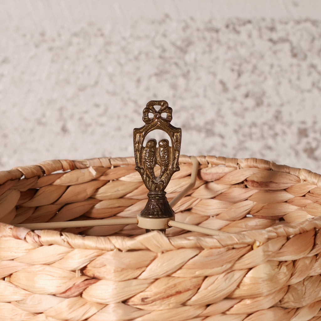 A close-up of a woven natural fiber basket with a decorative bronze-toned metal finial on top, set against a softly blurred light gray textured background.