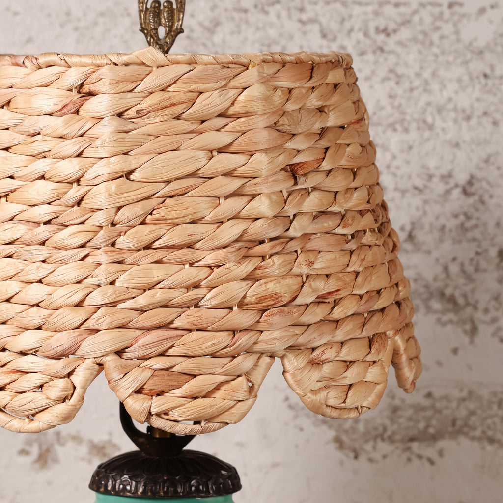 A close-up of a woven natural fiber lampshade, likely seagrass or raffia, with a scalloped edge and a dark metal finial at the top, set against a textured light gray wall.