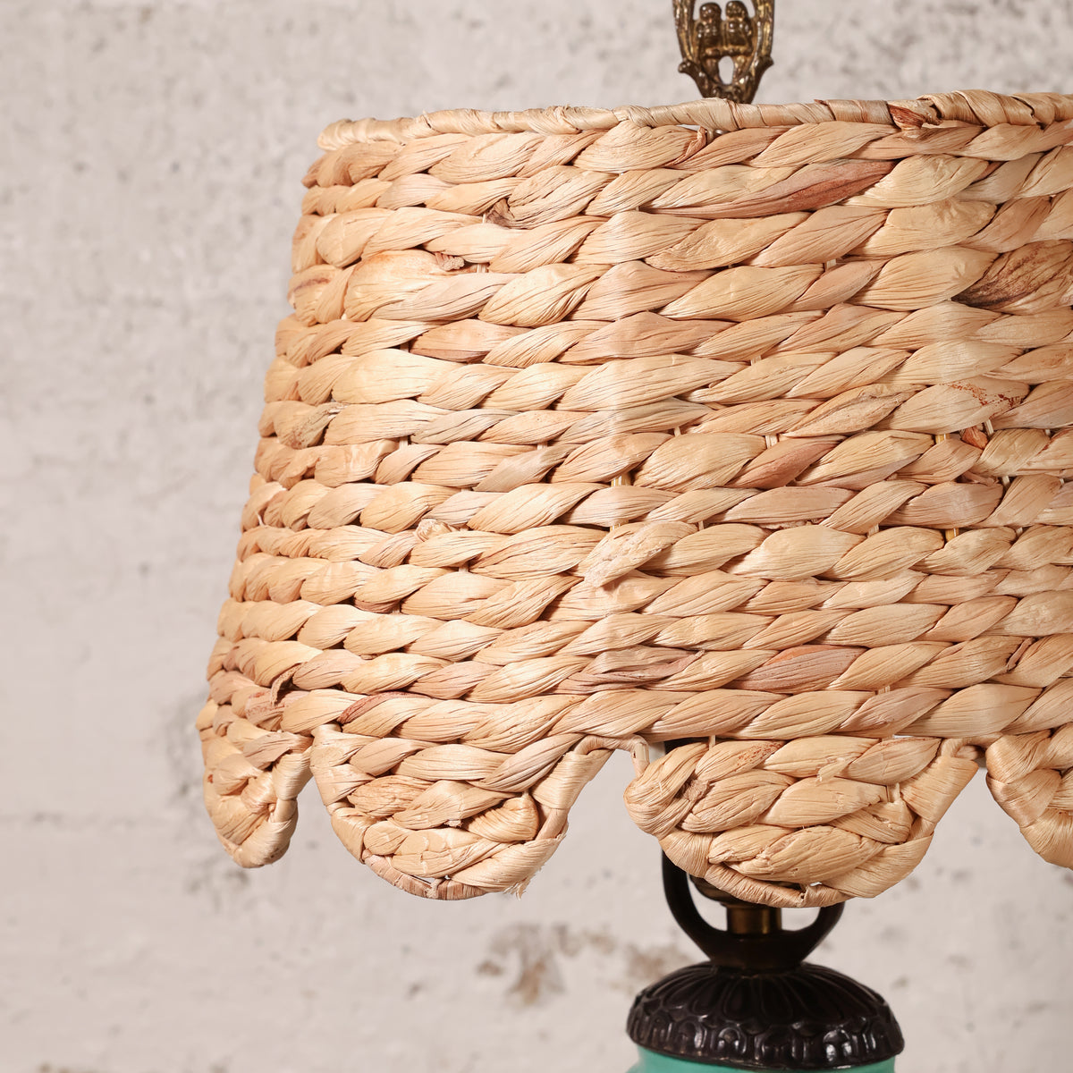 Close-up of a woven natural fiber lampshade in a warm tan color, attached to a dark metal lamp base against a textured off-white wall.