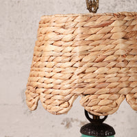 Close-up of a woven natural fiber lampshade in a warm tan color, attached to a dark metal lamp base against a textured off-white wall.