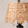 Close-up of a woven natural fiber lampshade in a warm tan color, attached to a dark metal lamp base against a textured off-white wall.