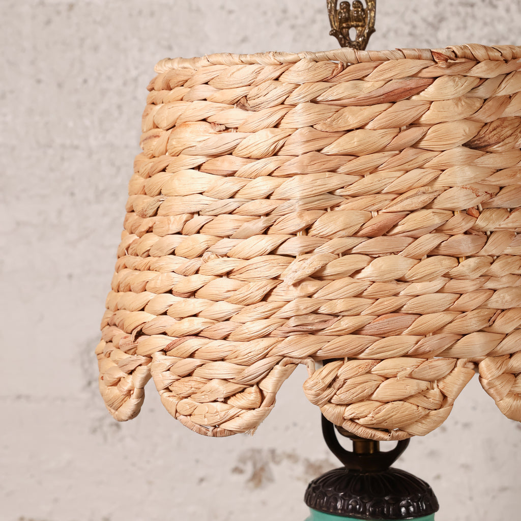 Close-up of a woven natural fiber lampshade in a warm tan color, attached to a dark metal lamp base against a textured off-white wall.