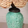 Close-up of a ceramic table lamp with a glossy turquoise-green relief base featuring a stylized figure design, topped with a dark metal collar and a woven natural-fiber shade.