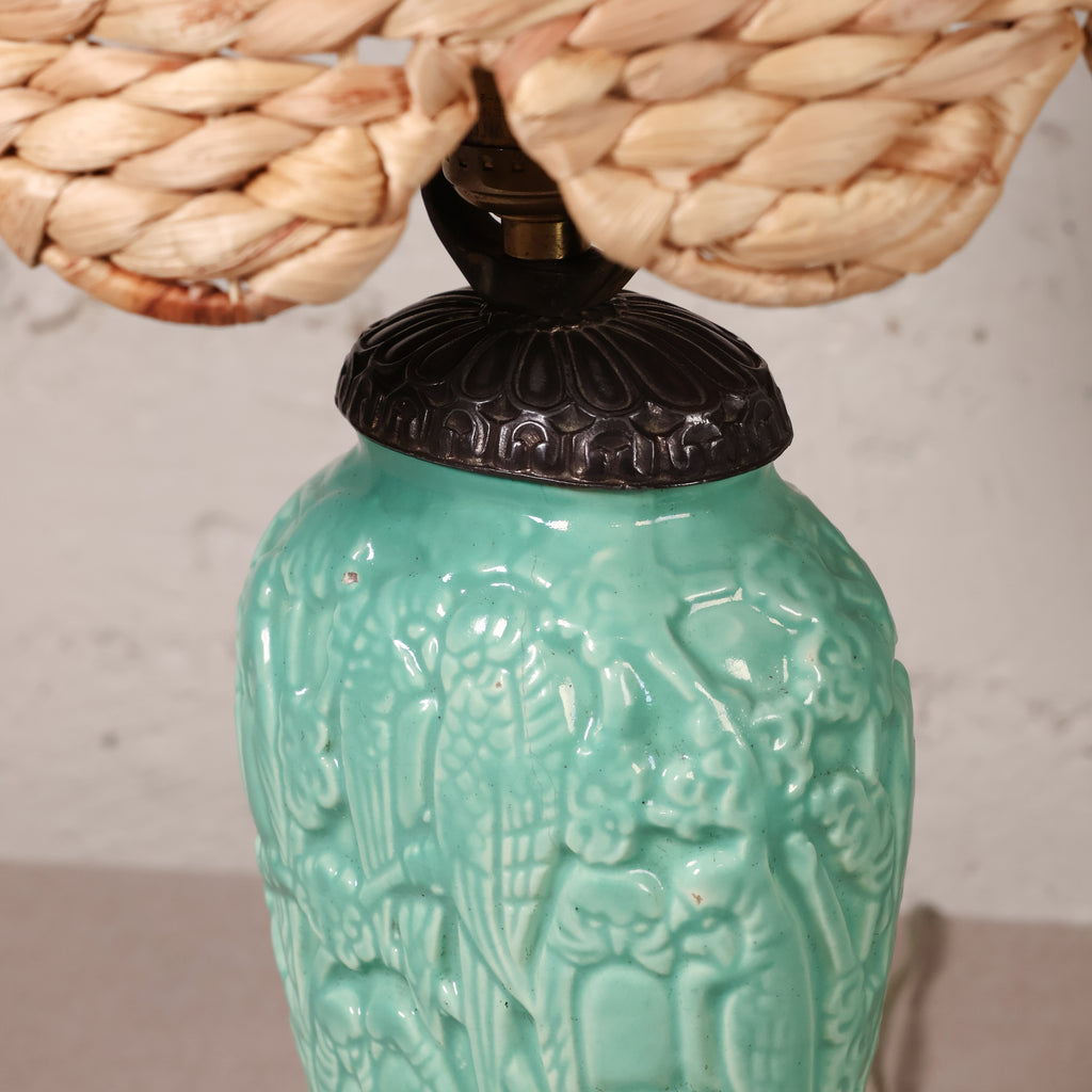 Close-up of a ceramic table lamp with a glossy turquoise-green relief base featuring a stylized figure design, topped with a dark metal collar and a woven natural-fiber shade.