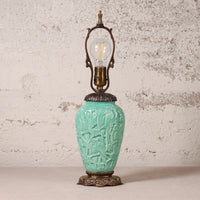 A vintage-style table lamp with a turquoise glazed ceramic vase-shaped base featuring embossed floral and bird motifs, topped with an ornate bronze metal frame and a clear filament light bulb, set against a textured off-white wall.