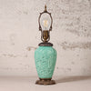 A vintage-style table lamp with a turquoise glazed ceramic vase-shaped base featuring embossed floral and bird motifs, topped with an ornate bronze metal frame and a clear filament light bulb, set against a textured off-white wall.