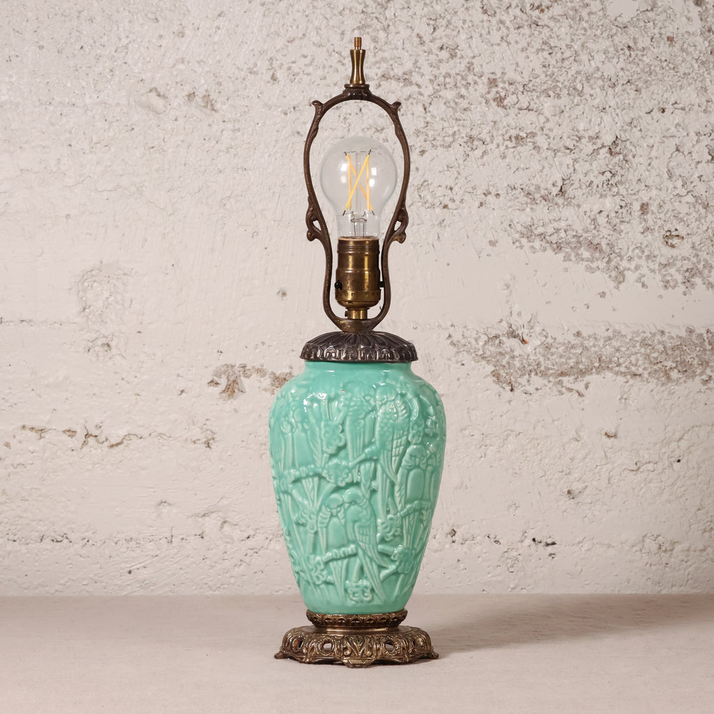 A vintage-style table lamp with a turquoise glazed ceramic vase-shaped base featuring embossed floral and bird motifs, topped with an ornate bronze metal frame and a clear filament light bulb, set against a textured off-white wall.