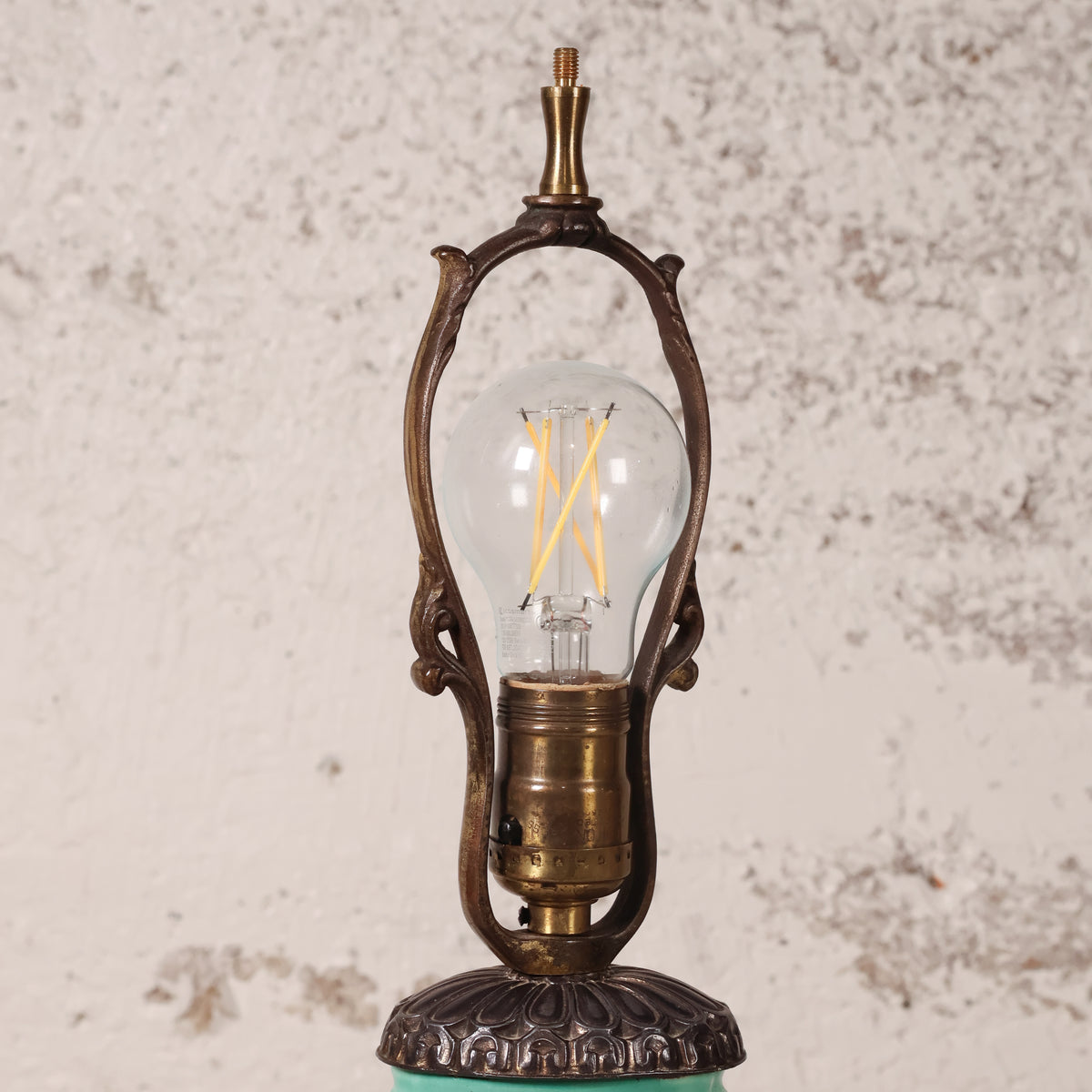 An ornate vintage-style table lamp with a clear glass globe bulb, featuring a bronze-finished metal frame and base, set against a textured off-white wall.