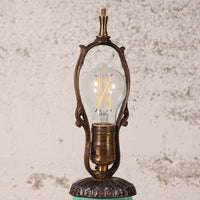 An ornate vintage-style table lamp with a clear glass globe bulb, featuring a bronze-finished metal frame and base, set against a textured off-white wall.