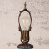 An ornate vintage-style table lamp with a clear glass globe bulb, featuring a bronze-finished metal frame and base, set against a textured off-white wall.