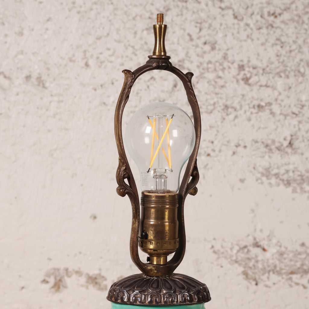 An ornate vintage-style table lamp with a clear glass globe bulb, featuring a bronze-finished metal frame and base, set against a textured off-white wall.