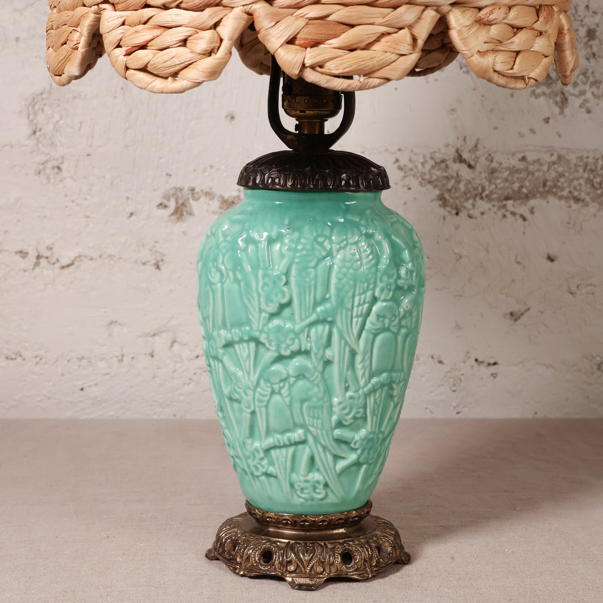 A vintage-style table lamp with a turquoise glazed ceramic vase base featuring embossed floral and bird motifs, a dark metal neck, and a woven natural-fiber shade visible at the top.