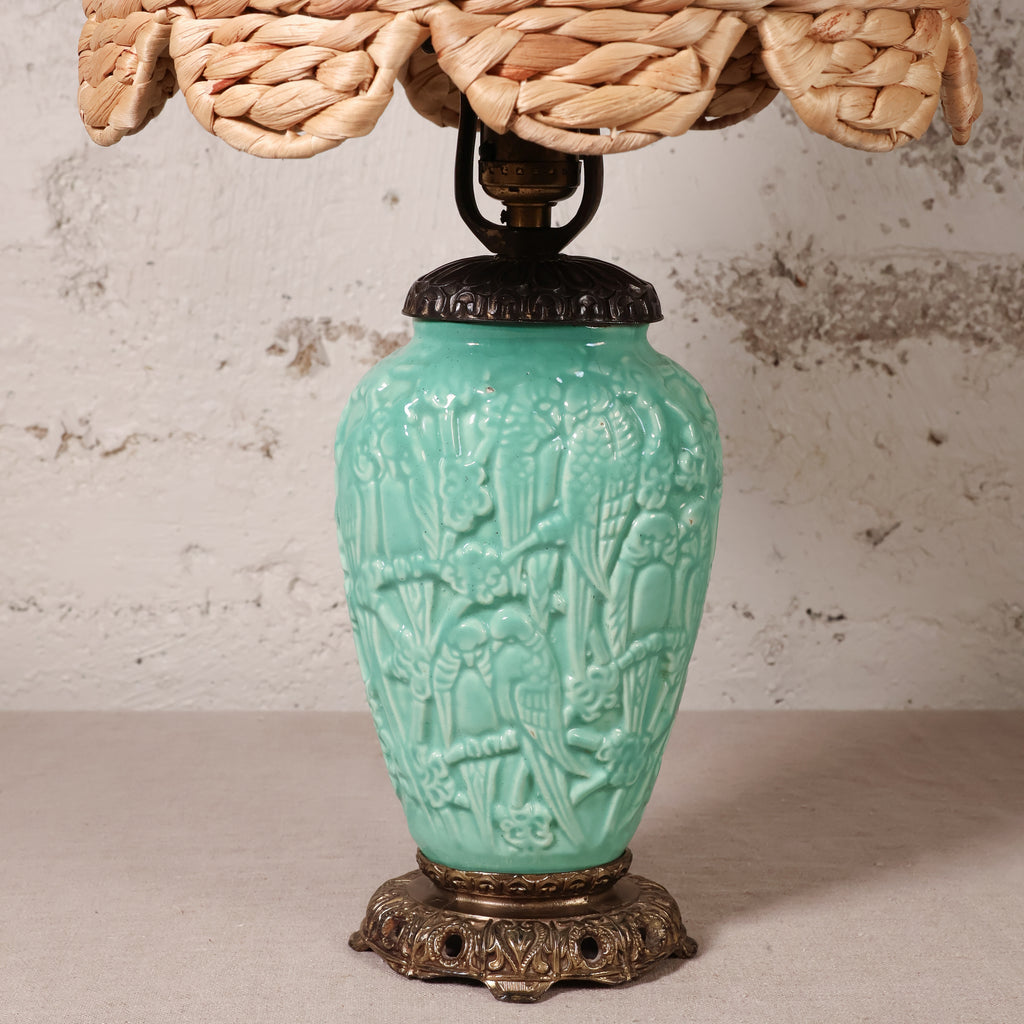 A vintage-style table lamp with a turquoise glazed ceramic vase base featuring embossed floral and bird motifs, a dark metal neck, and a woven natural-fiber shade visible at the top.
