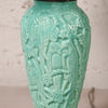 A close-up of a turquoise glazed ceramic vase featuring embossed relief carvings of birds and floral motifs, set against a light textured wall.