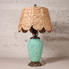 A table lamp with a natural woven straw shade and a turquoise ceramic base featuring a detailed floral relief, set against a rustic, light gray textured wall.