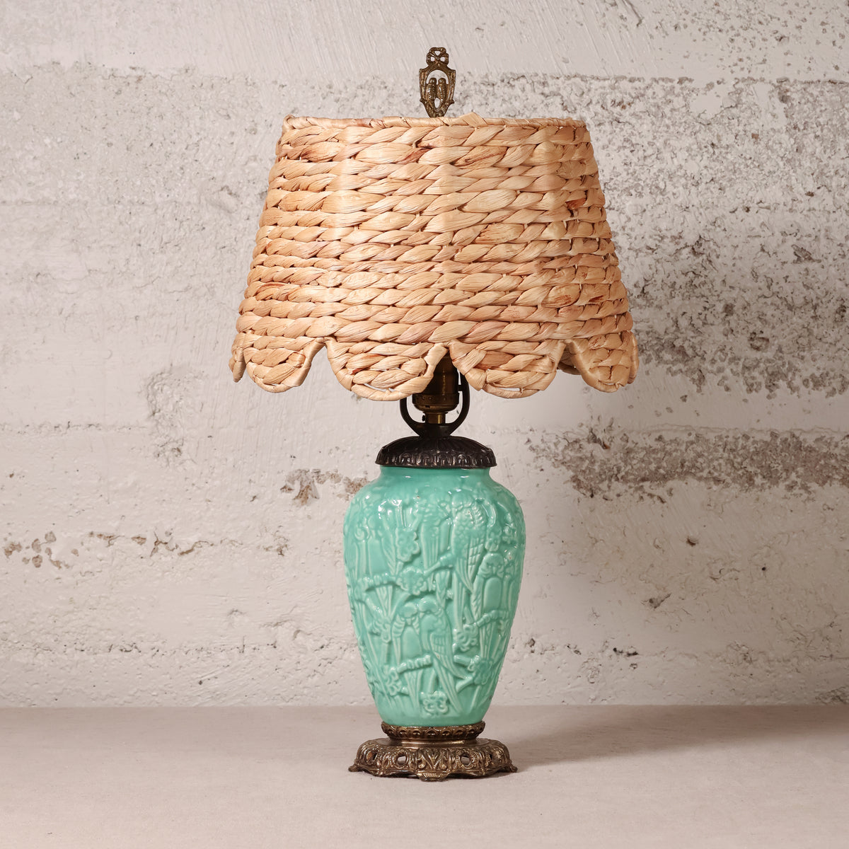 A table lamp with a natural woven straw shade and a turquoise glazed ceramic base featuring embossed floral details, set against a textured light gray wall.