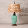 A table lamp with a natural woven straw shade and a turquoise glazed ceramic base featuring embossed floral details, set against a textured light gray wall.
