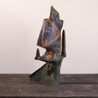 A rustic, handcrafted ceramic sculpture of an abstract figure, featuring weathered brown and green patina textures, displayed on a dark wooden surface against a plain off-white wall.