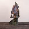 A rustic, handcrafted ceramic sculpture of an abstract figure, featuring weathered brown and green patina textures, displayed on a dark wooden surface against a plain off-white wall.