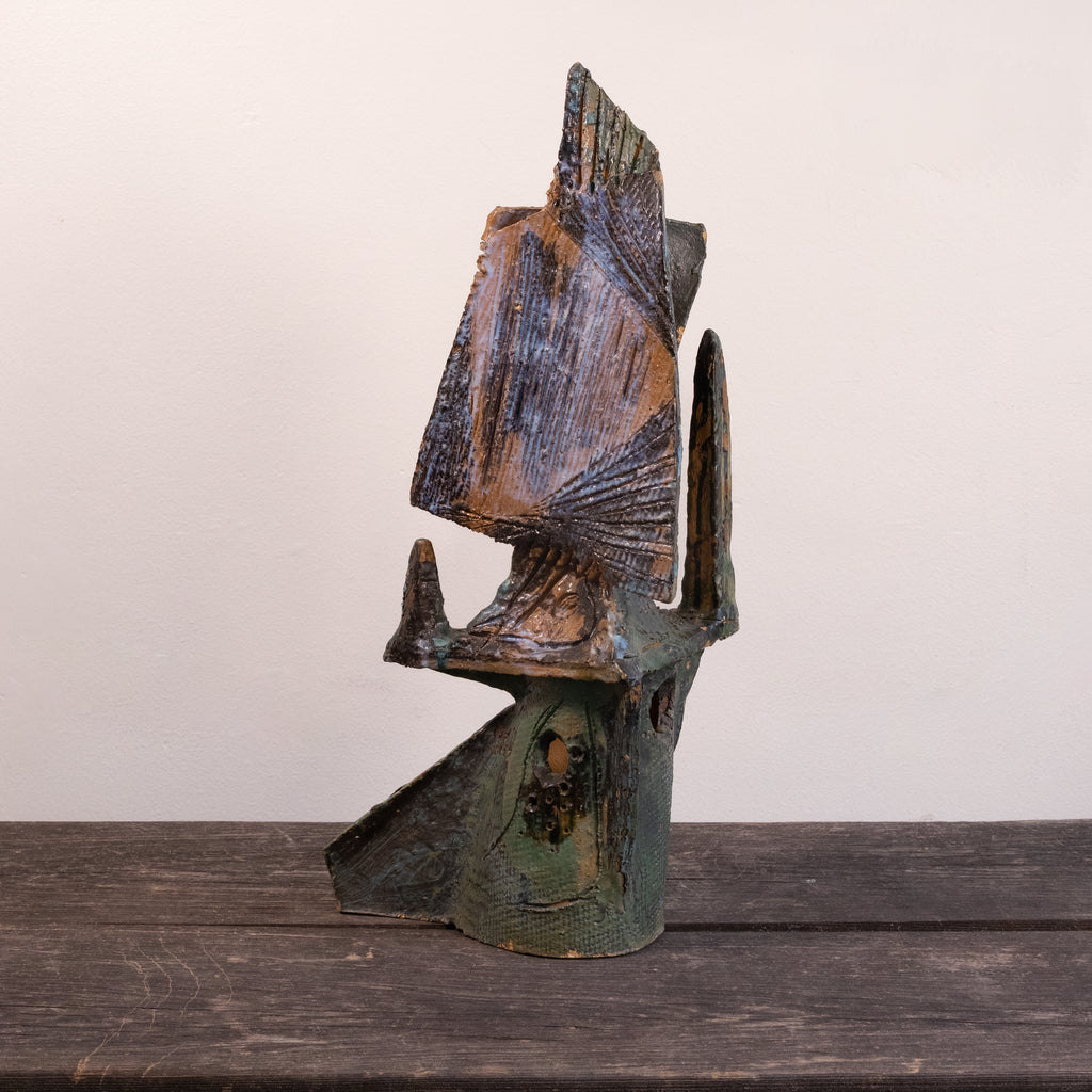 A rustic, handcrafted ceramic sculpture of an abstract figure, featuring weathered brown and green patina textures, displayed on a dark wooden surface against a plain off-white wall.