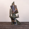 An abstract sculptural art piece with a textured, weathered finish in earthy tones of brown, green, and blue, displayed on a dark wooden surface against a plain off-white wall.
